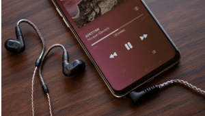 _Black Sennheiser IE 200 in-ear headphones placed next to a smartphone displaying a music player interface on a wooden surface. The screen shows a track titled 'APETITAN' with playback controls.
