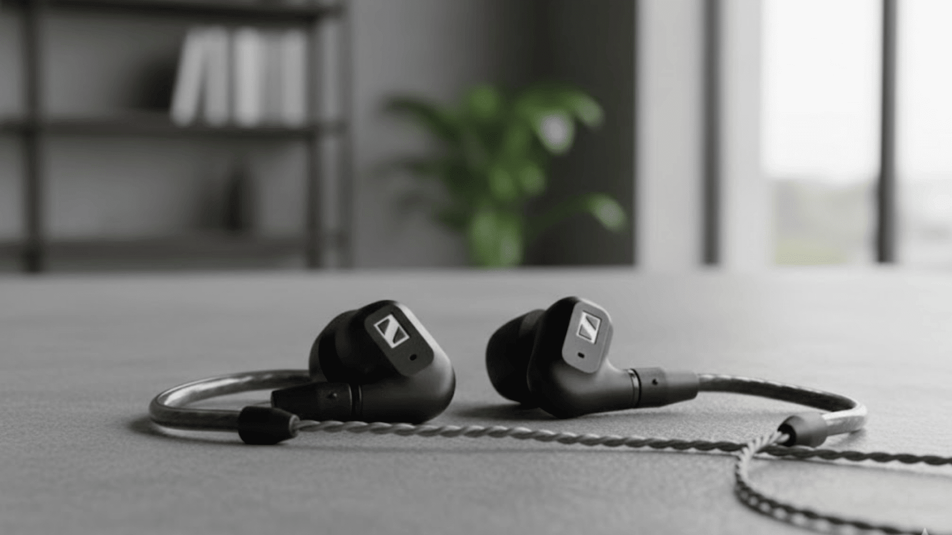 _Two Sennheiser IE 200 in-ear headphones displayed on a gray surface with a blurred background of a modern room featuring greenery and bookshelves. This image supports a Sennheiser IE 200 Review.