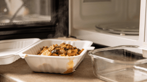 Takeout container with food placed inside an open microwave next to a glass dish