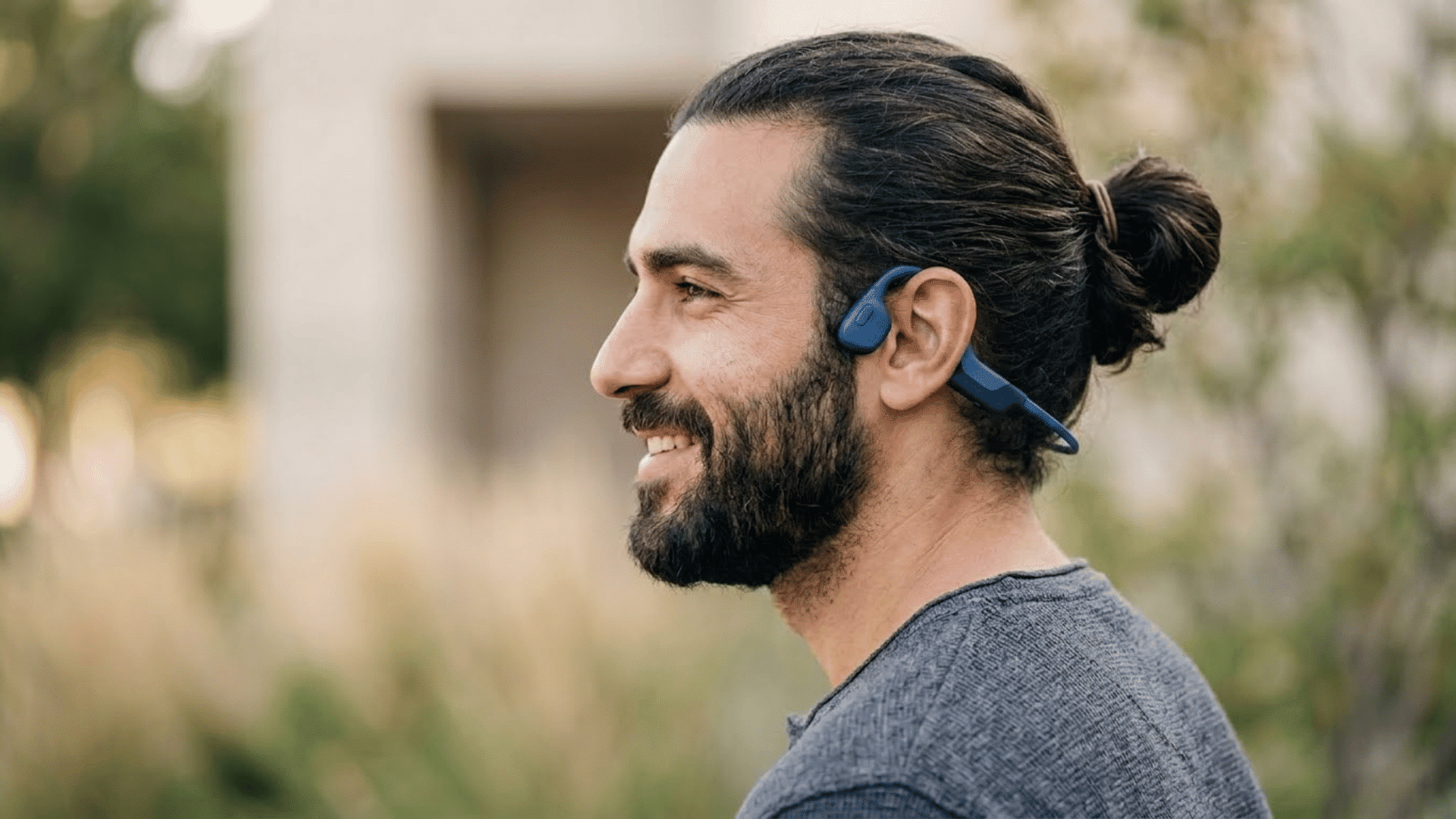 Man wearing blue bone conduction headphones while outdoors with hair tied in a bun.
