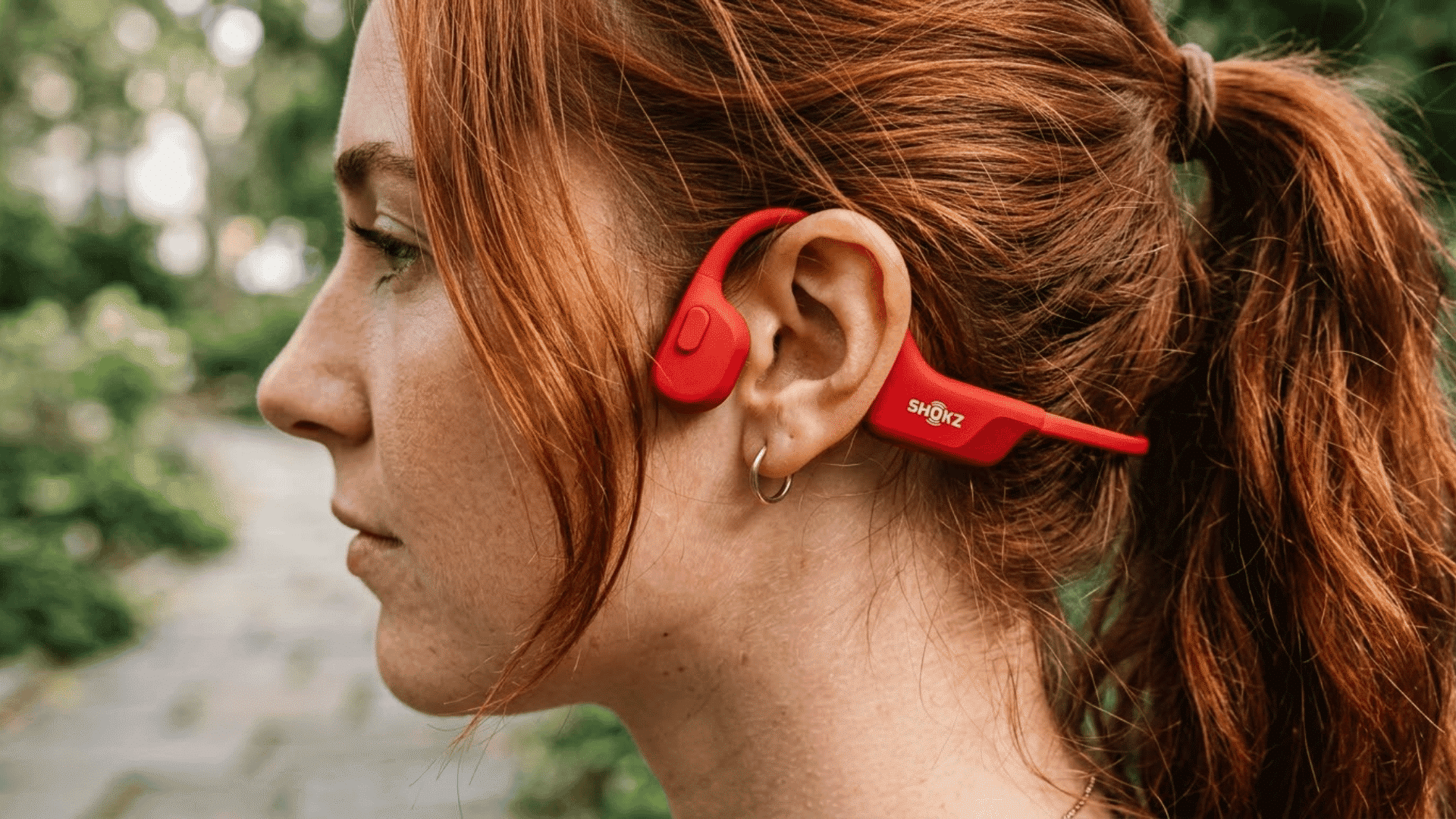 Person wearing red Shokz bone conduction headphones while outdoors on a nature trail.